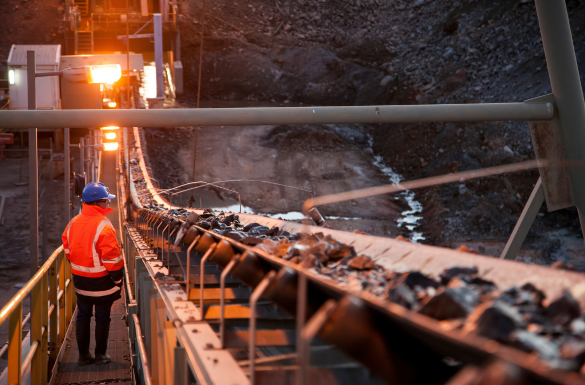 A person in a safety vest and helmet observes an outdoor mining conveyor belt transporting raw materials. The scene is lit by industrial lighting, highlighting the machinery and rocky terrain. The sky is dim, suggesting early morning or late evening. A person in a safety vest and helmet observes an outdoor mining conveyor belt transporting raw materials. The scene is lit by industrial lighting, highlighting the machinery and rocky terrain. The sky is dim, suggesting early morning or late evening.